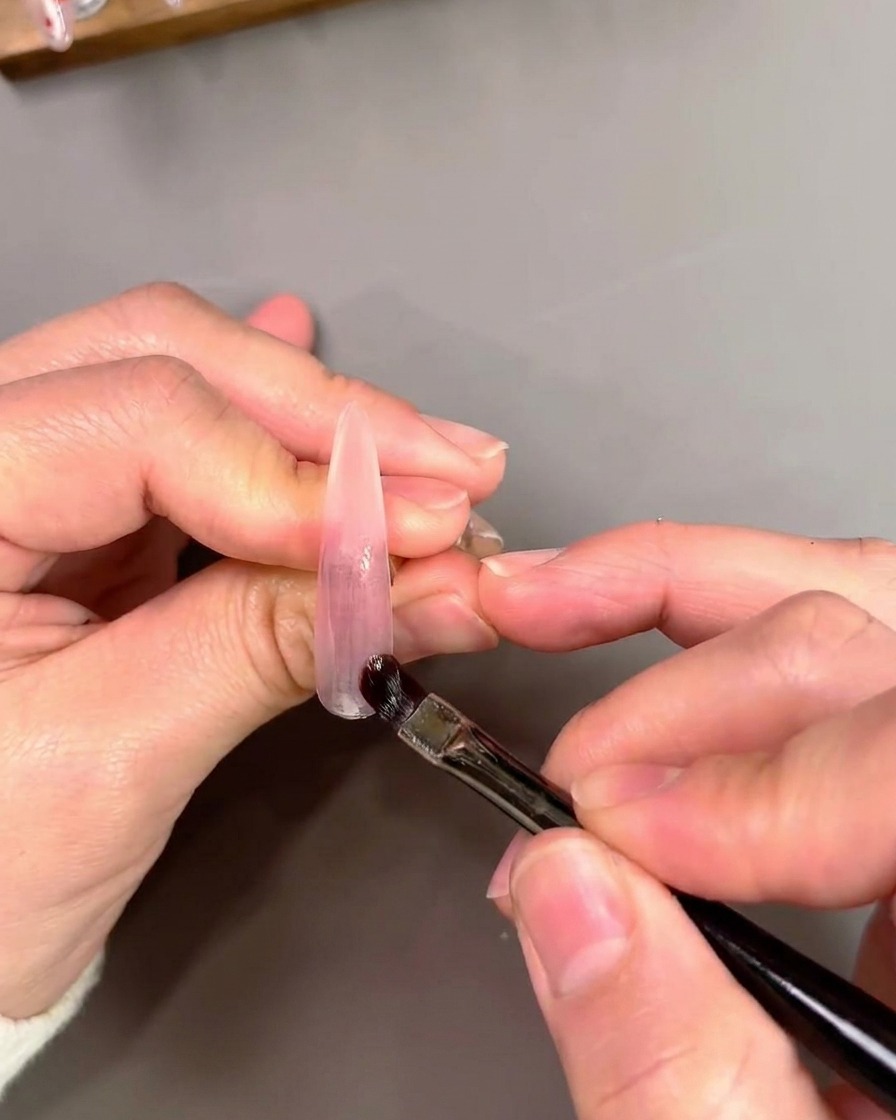 Un pincel plano aplicando y difuminando esmalte gel morado cat-eye sobre una uña artificial.