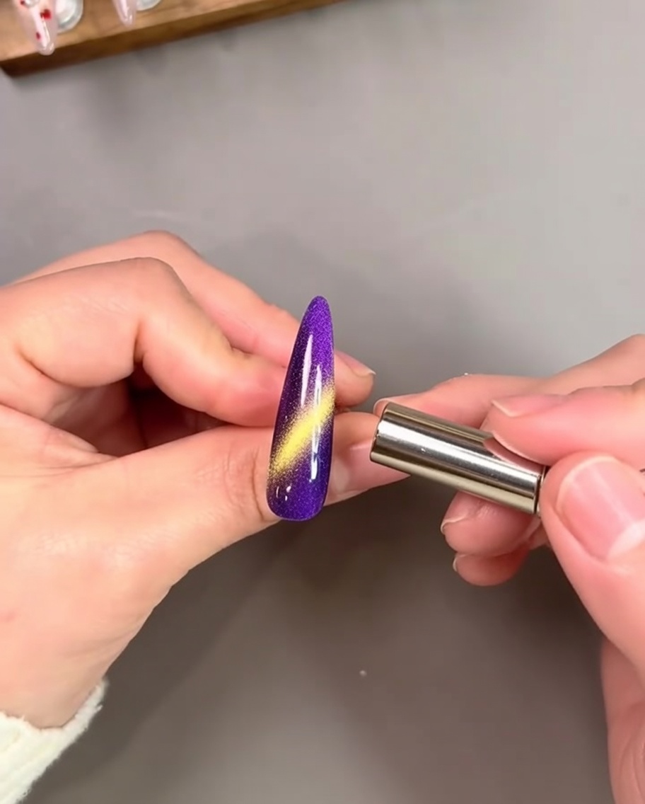 Una mano sosteniendo un imán cilíndrico cerca de una uña morada vibrante, activando un destello dorado intenso.