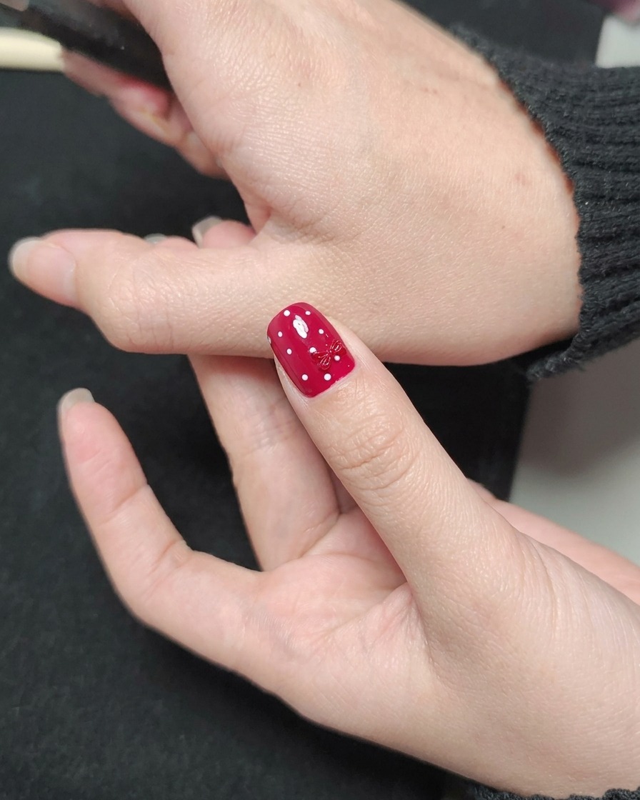 Un pincel de esmalte aplicando un top coat transparente brillante sobre una uña roja con lunares blancos y un dije de lazo rojo.