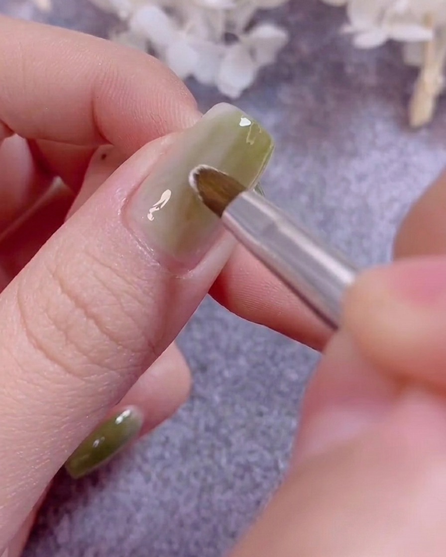 Un pincel fino de decoración pintando delicados pétalos de flores blancas sobre una uña recubierta de gel verde.