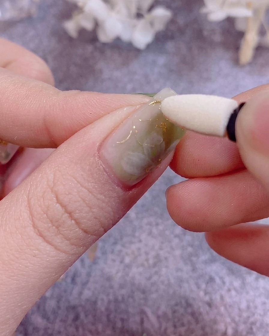 Primer plano de pinzas aplicando delicadas escamas de pan de oro sobre una uña pintada.
