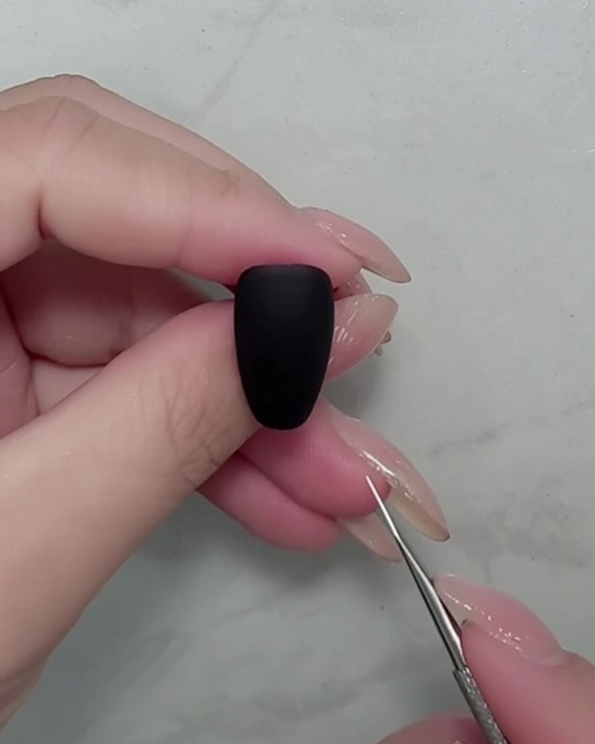 Primer plano de un pincel de uñas pintando tallos blancos finos sobre una uña negra mate.