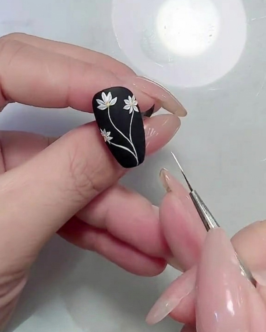 Primer plano de un pincel de detalle aplicando formas de hoja verdes junto a los tallos de margarita en una uña negra mate.