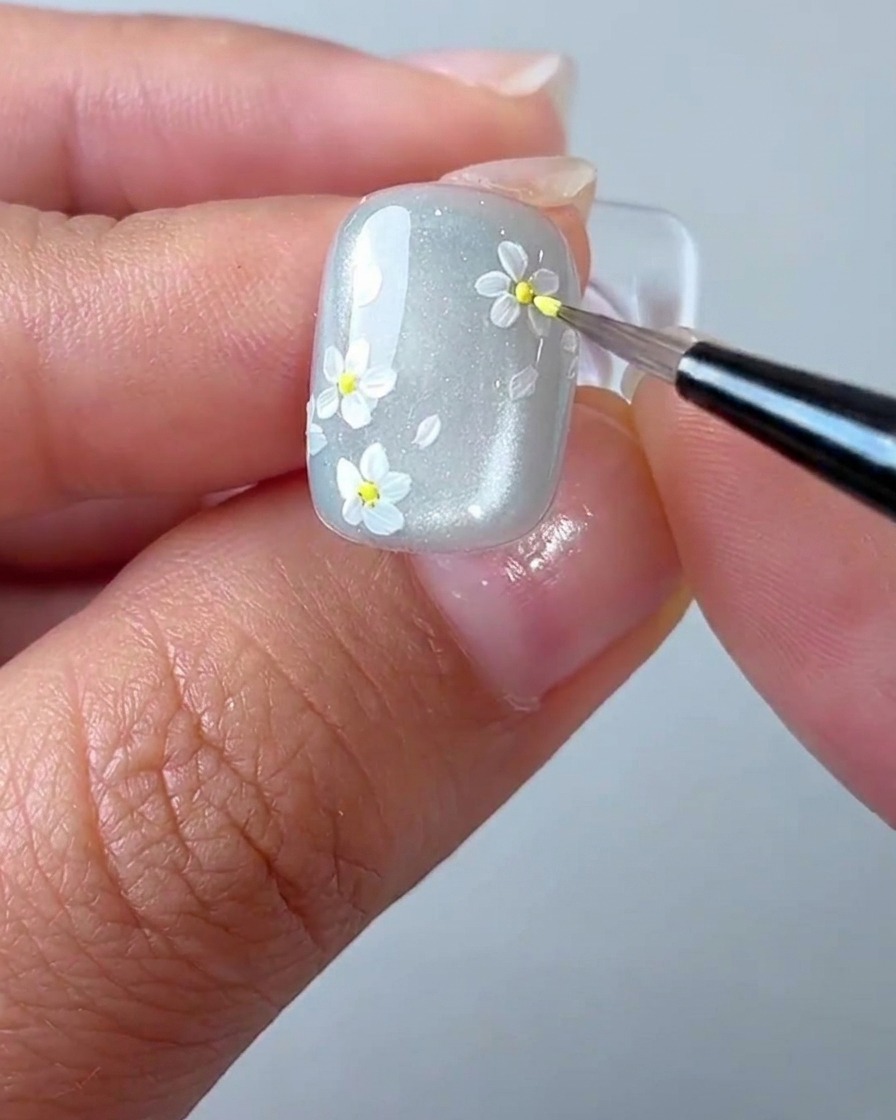 Un pincel de detalle aplicando un pequeño punto amarillo en el centro de una margarita blanca en la uña.