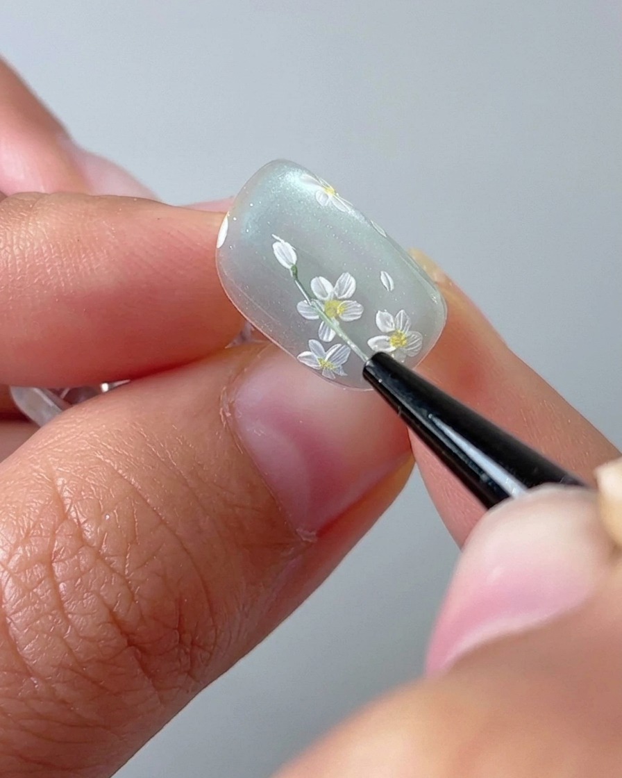 Un primer plano de un pincel de uñas dibujando un tallo verde fino sobre una base de gel cat-eye.