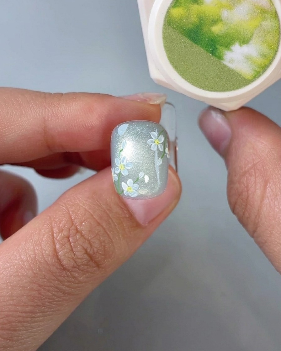 Una vista de primer plano de un pincel aplicando pequeños detalles de hojas verdes sobre una uña con arte de margarita.