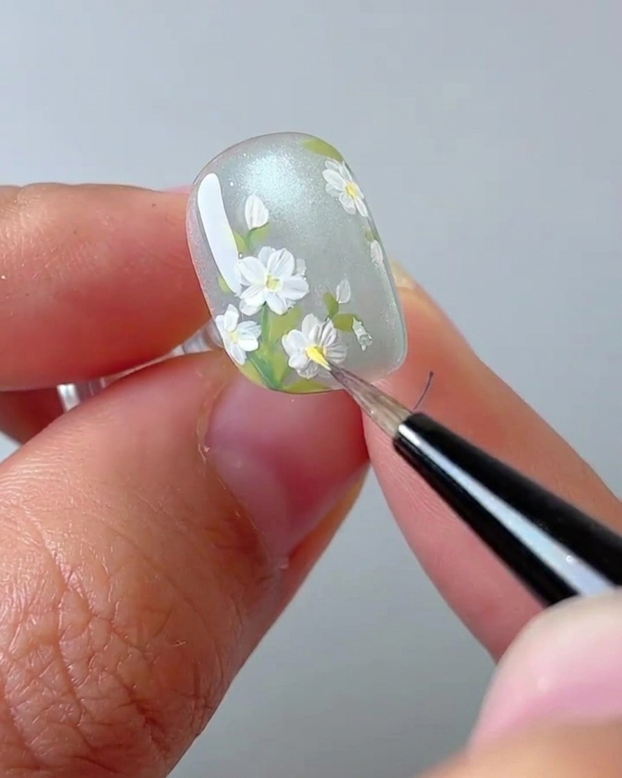 Una toma de primer plano de una artista de uñas usando un pincel para colocar un punto amarillo en el centro de una margarita blanca.