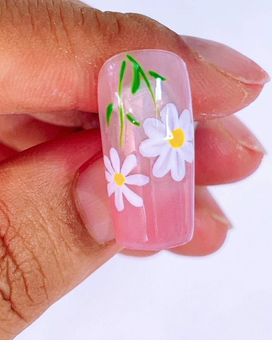 Primer plano de una uña con degradado rosa siendo pintada con una segunda margarita más pequeña usando gel blanco y un pincel liner fino.
