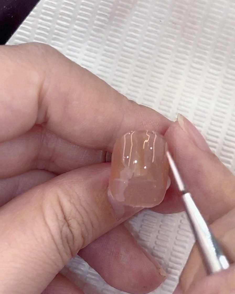 Un primer plano de un pincel liner aplicando un pequeño punto guía blanco en el centro de una uña de gel nude.