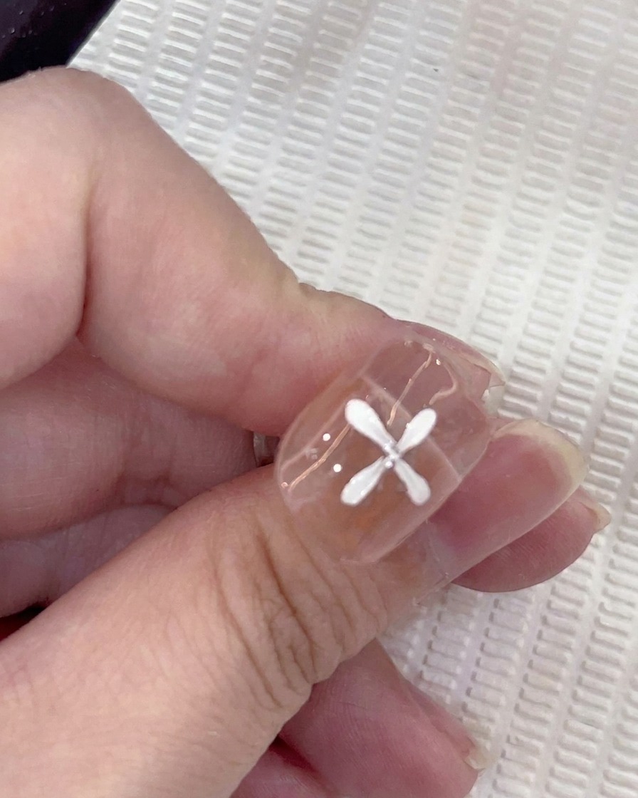 Un primer plano de una uña con cinco pétalos blancos pintados en un patrón circular, con un pincel añadiendo un sexto pétalo.