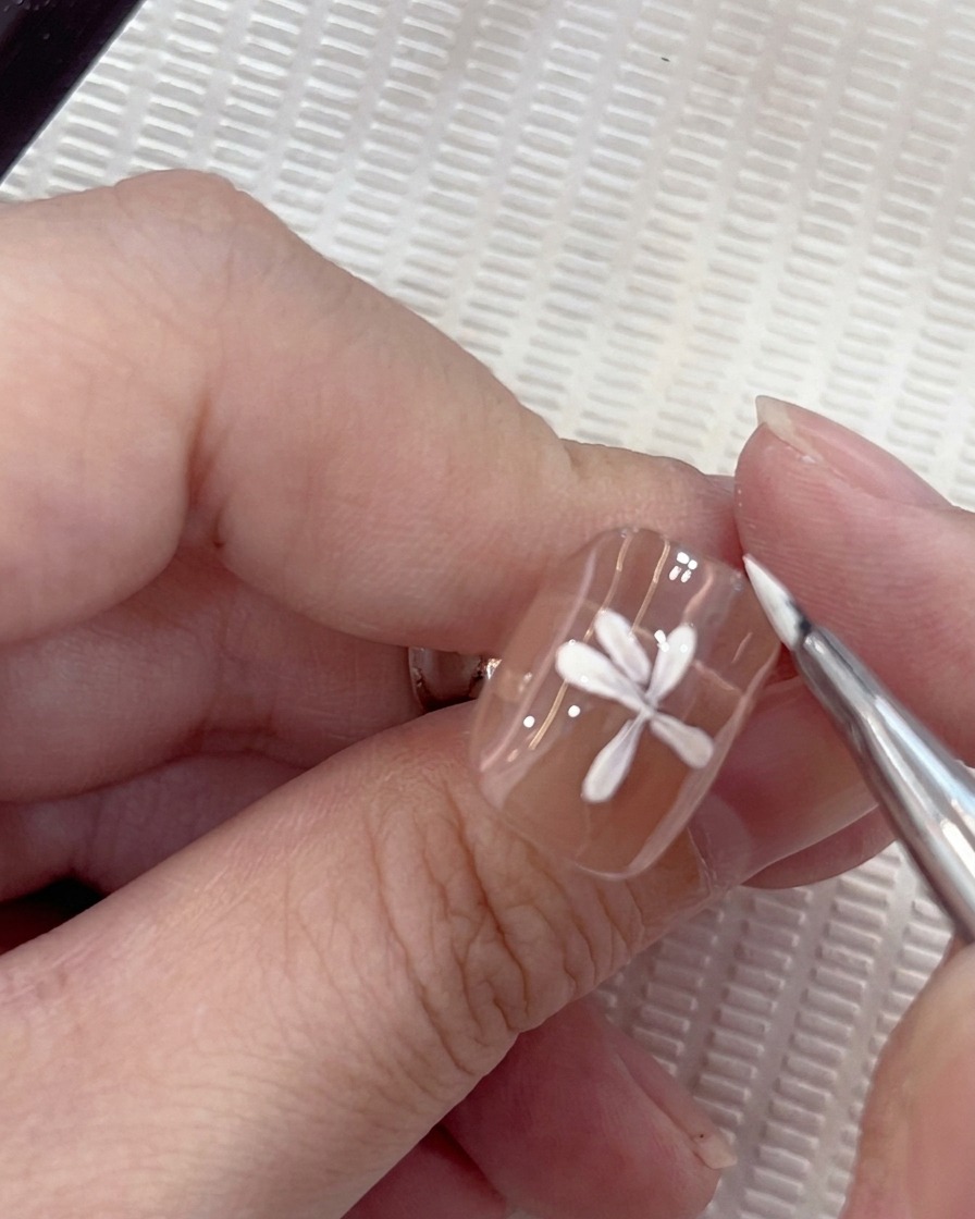 Un primer plano de un diseño de arte de uñas de margarita blanca donde un séptimo pétalo está siendo colocado cuidadosamente por un pincel redondo.