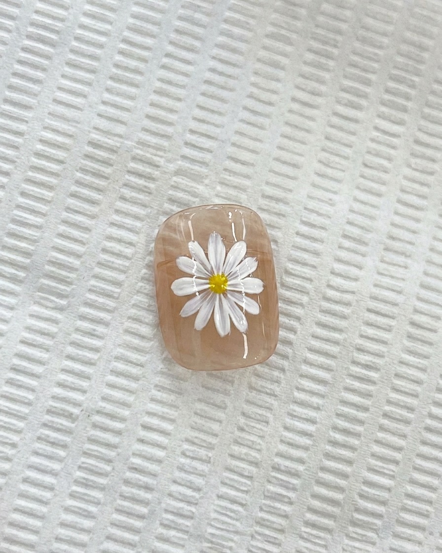 Un primer plano de una margarita blanca en una uña, con un pincel de detalle fino aplicando un centro amarillo.