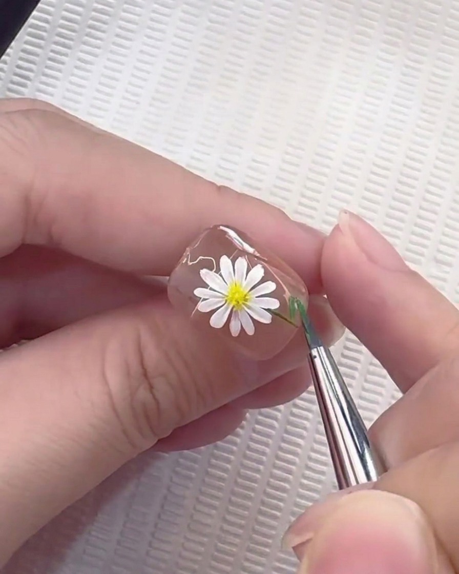 Un pincel liner tirando de una línea verde fina para formar un tallo en una uña decorada con margaritas.