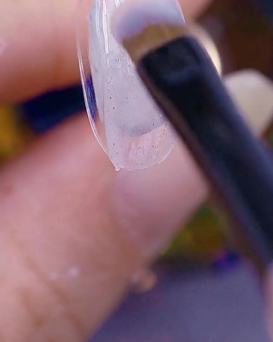 Primer plano de un pincel aplicando un gel texturizado transparente sobre la punta de una uña natural.