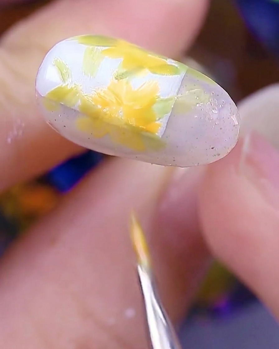 Primer plano de la uña mostrando varias margaritas amarillo-naranja pintadas sobre las hojas, mientras el pincel termina los últimos pétalos.