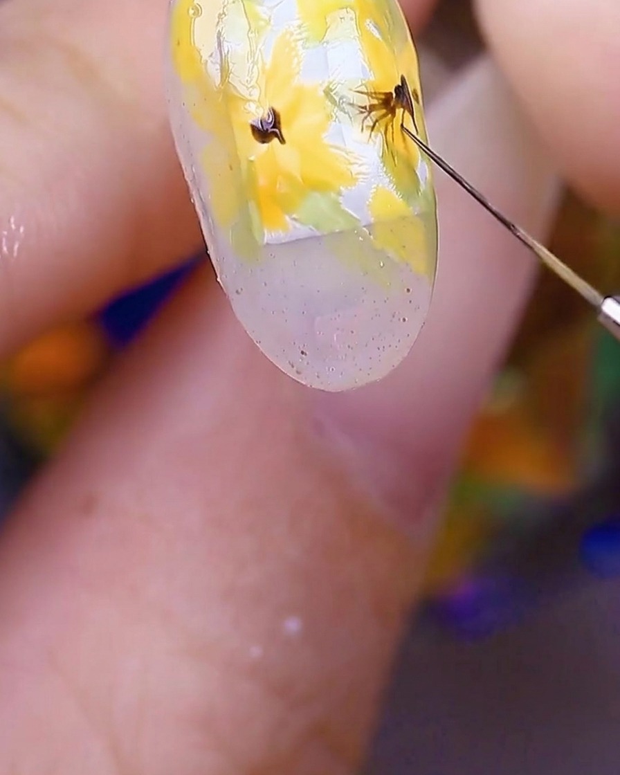 Primer plano del nail art mostrando un pincel trazando líneas finas de color marrón oscuro desde el centro de las flores amarillas hacia afuera.