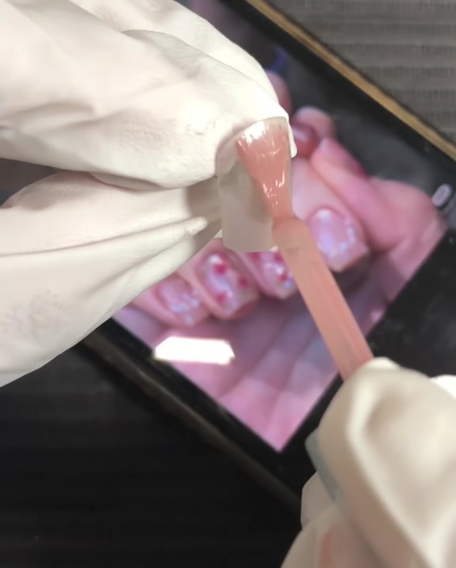 Pincel aplicando esmalte de gel rosa nude en toda la superficie de la uña.