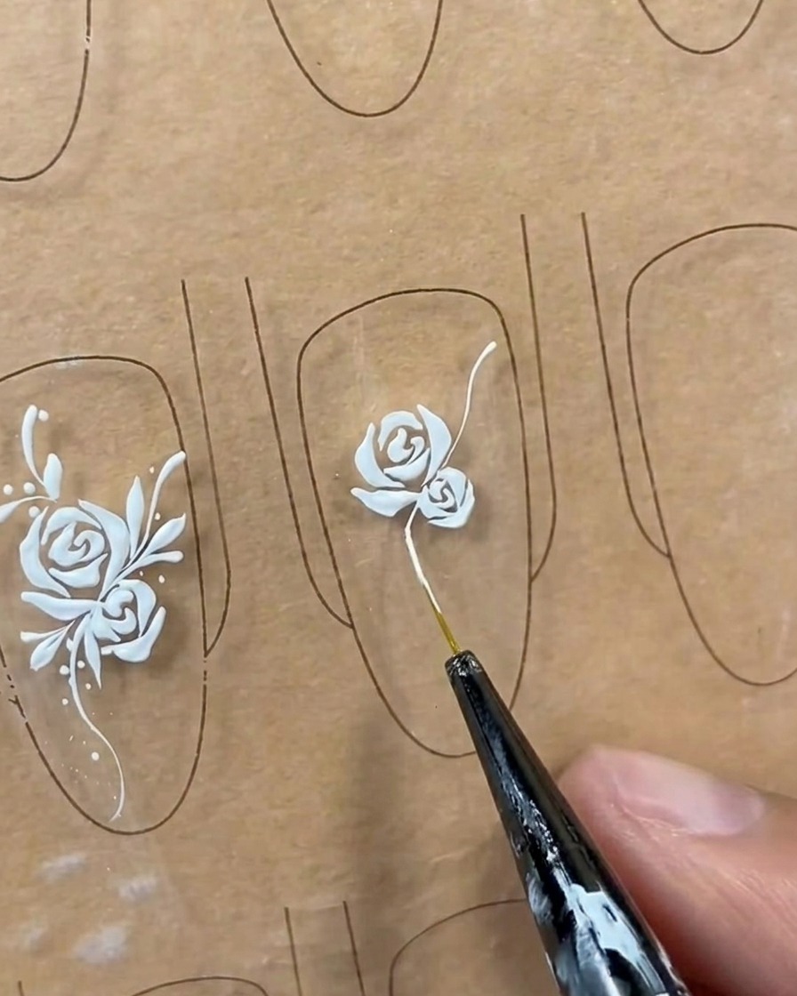 Una línea de enredadera blanca larga y fluida siendo dibujada desde la base de un diseño de rosa en un tip de uña usando un pincel liner.