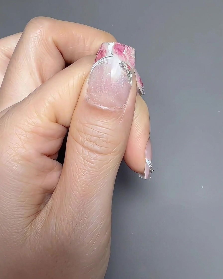 Un pincel de uñas aplicando un top coat transparente y brillante sobre una uña decorada con motivos florales.