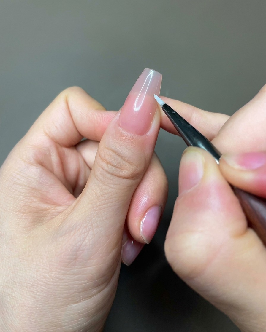 Primer plano de un pincel liner aplicando gel blanco para crear formas de pétalos sobre una uña de color nude.