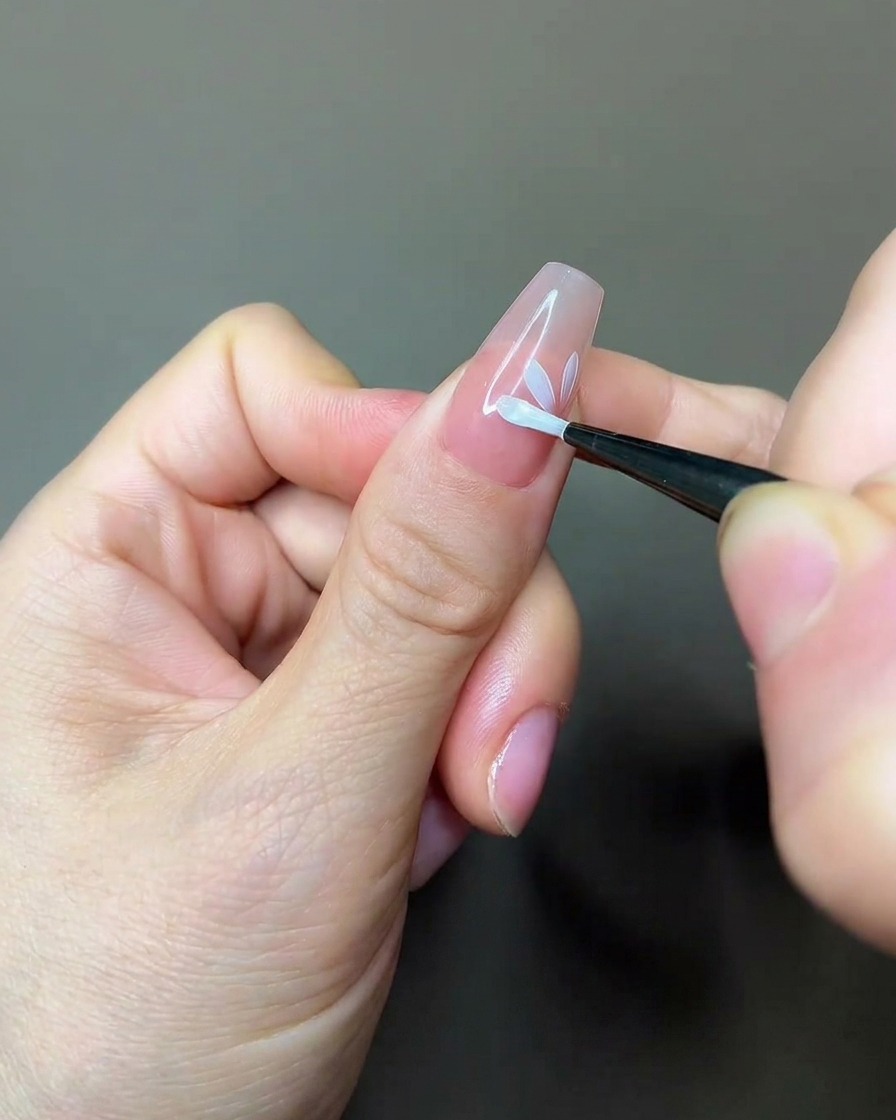 Un pincel de uñas aplicando una capa de top coat mate sobre un diseño floral parcialmente pintado en una uña nude.
