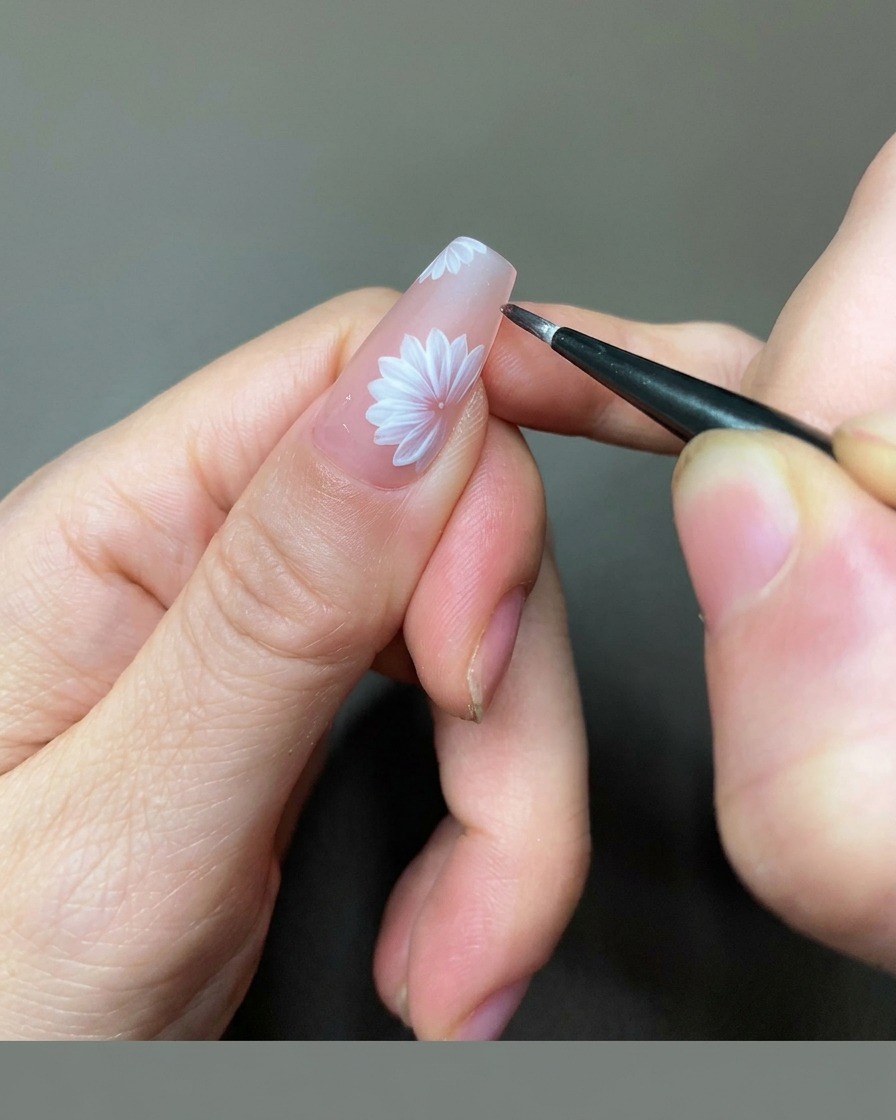 Vista en primer plano de una técnica de uñas usando un pincel liner fino para añadir detalles de gel dorado al centro de una margarita blanca en una uña nude.