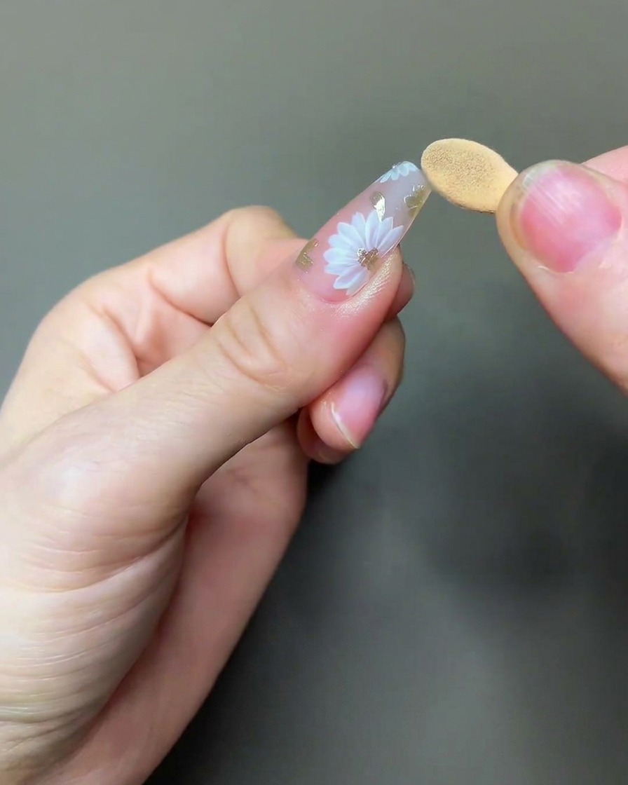 Un pincel aplicando un top coat transparente y brillante sobre el diseño de uñas de margarita y cromo completado.