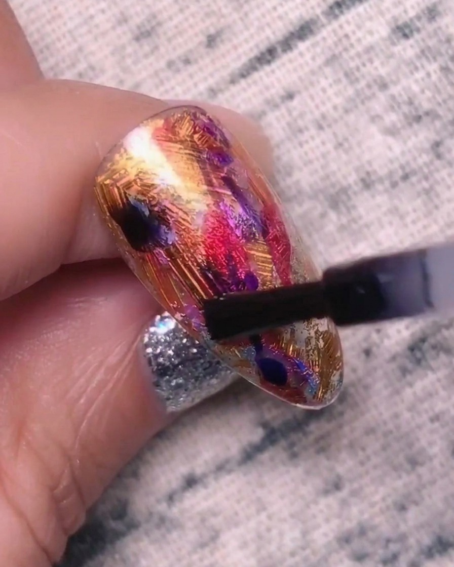 Primer plano de una uña siendo punteada con gel azul-púrpura, mostrando los colores comenzando a mezclarse y expandirse en la capa base.