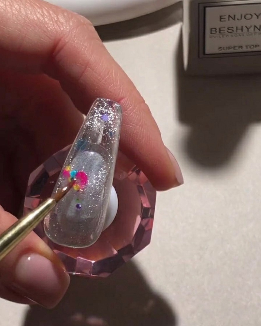 Un pincel de detalle colocando lentejuelas redondas y coloridas sobre una superficie de uña brillante.