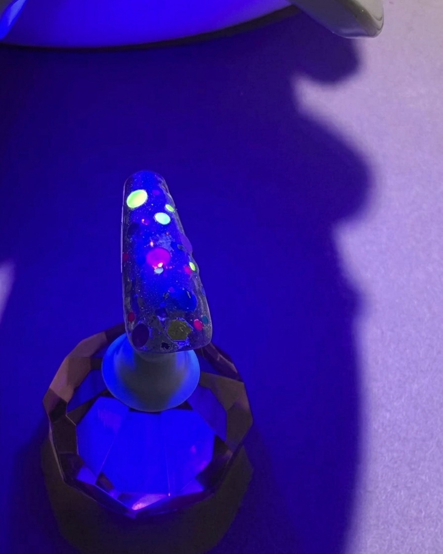 Una uña con lentejuelas coloridas bajo la luz violeta de una lámpara UV durante el proceso de curado.