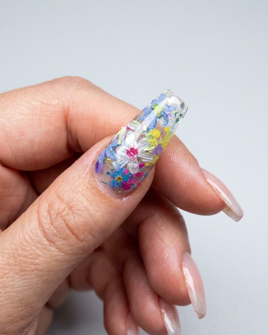 Primer plano de un pincel aplicando pétalos florales 3D multicolores sobre una uña decorada.