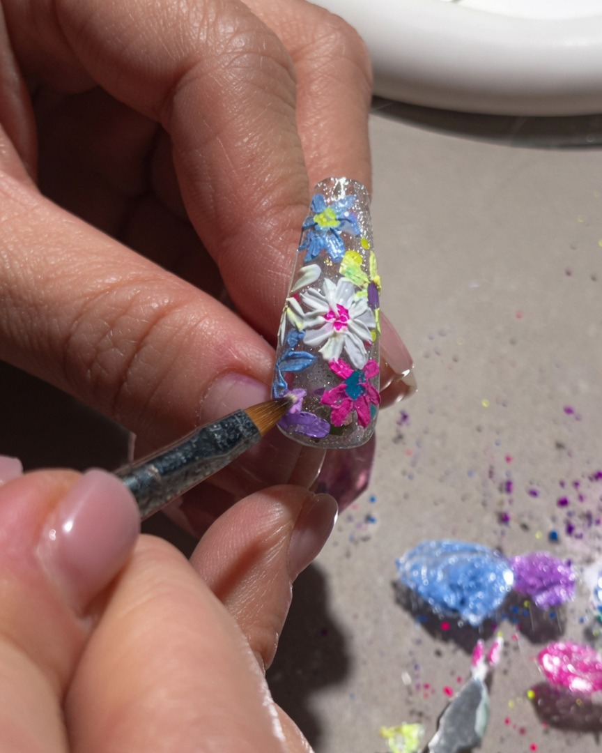 Primer plano de un pincel fino añadiendo luces blancas y detalles centrales a un diseño de uñas floral en 3D.