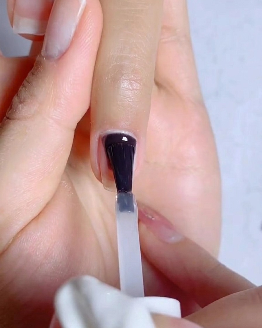 Primer plano de un pincel aplicando una capa transparente y fina de base coat de gel sobre una uña natural preparada.