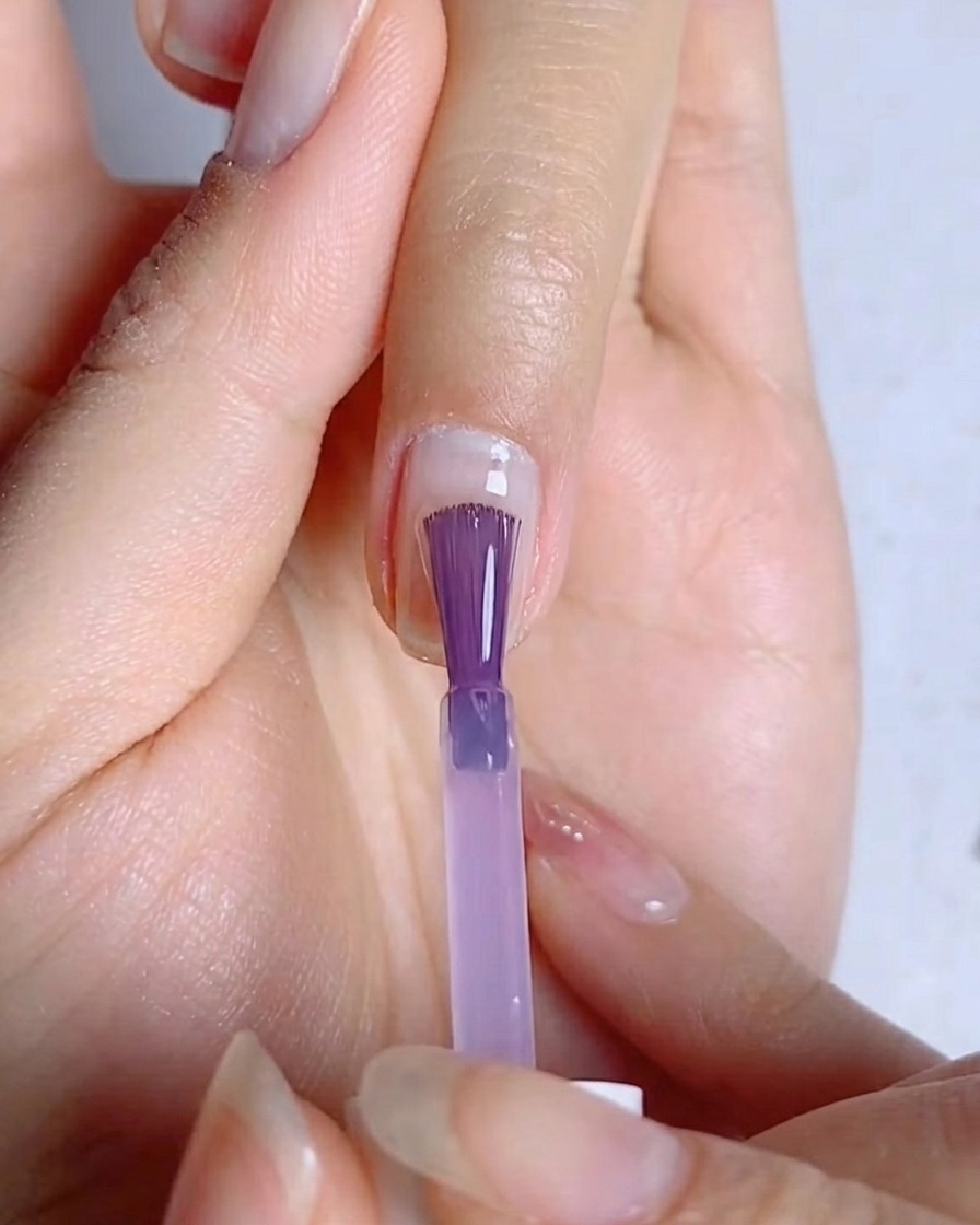 Un pincel de uñas extendiendo una capa fina y uniforme de esmalte gel azul-morado sobre la superficie de la uña.