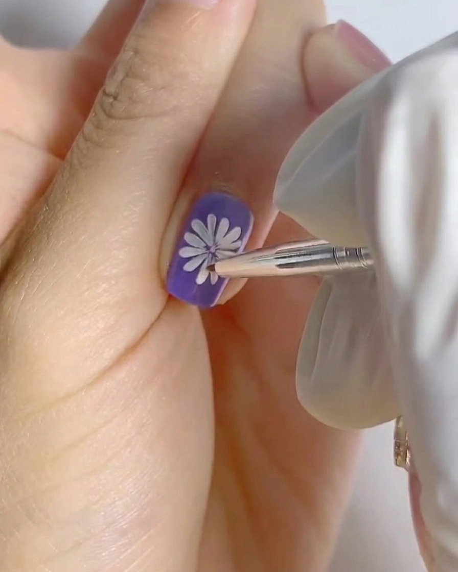 Un dedo con el arte de margarita blanca completado sobre una base morada colocado bajo una pequeña lámpara de uñas profesional para curar.