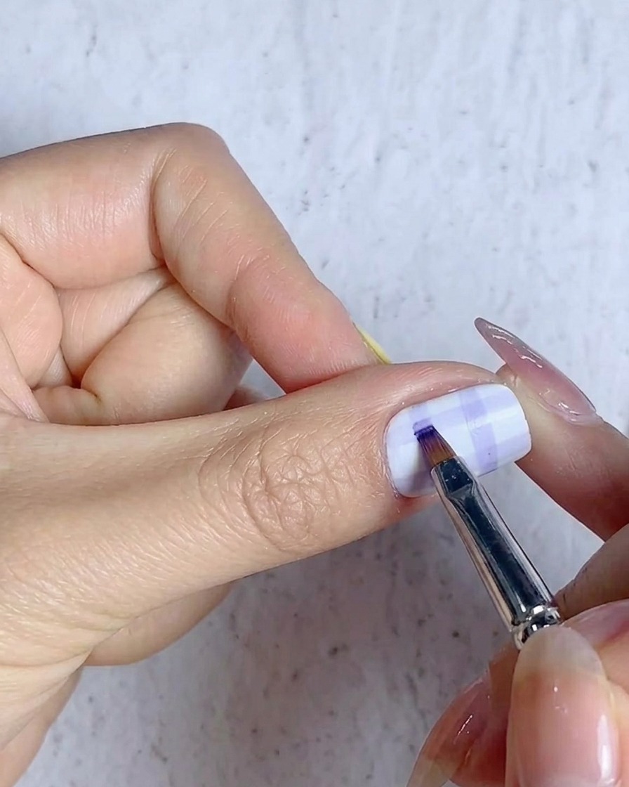 Un pincel fino rellenando las intersecciones de la cuadrícula en un diseño de uña a cuadros con gel azul.