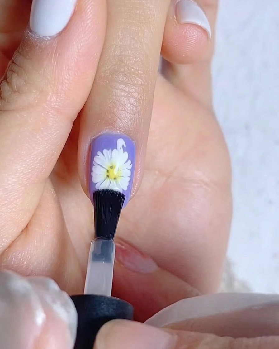 Un pincel aplicando una capa de top coat mate sobre una uña morada que presenta un diseño de margarita blanca.