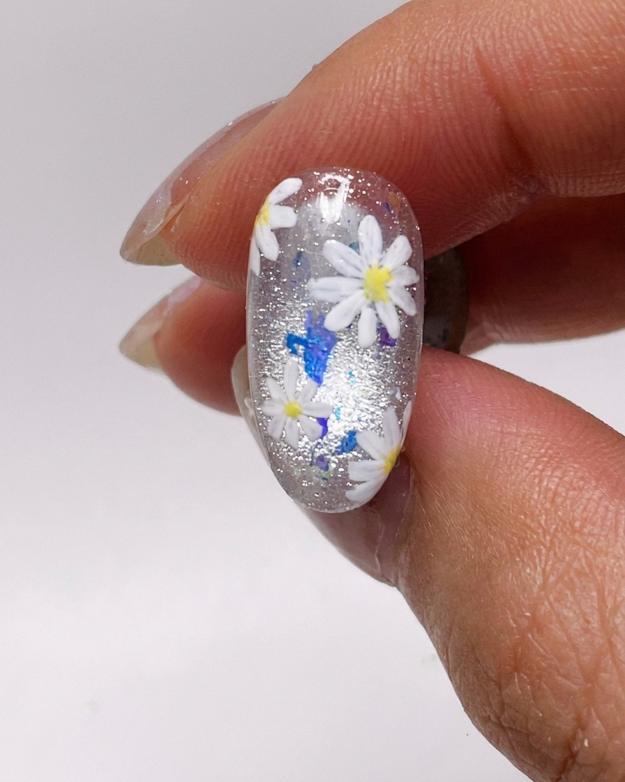 Primer plano de un pincel de uñas aplicando una capa fina de esmalte de gel plateado cat-eye en una uña natural.