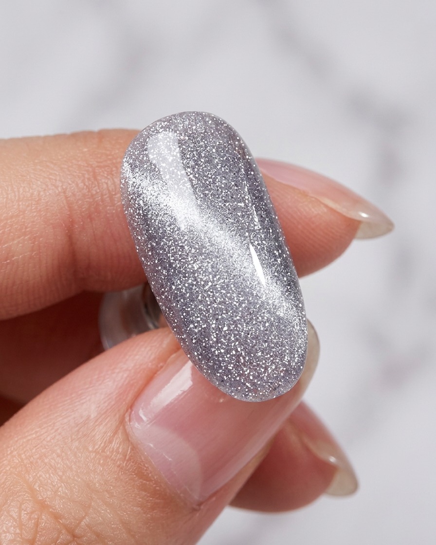 Una herramienta magnética siendo sostenida cerca de una uña con esmalte plateado cat-eye húmedo, revelando una línea brillante.