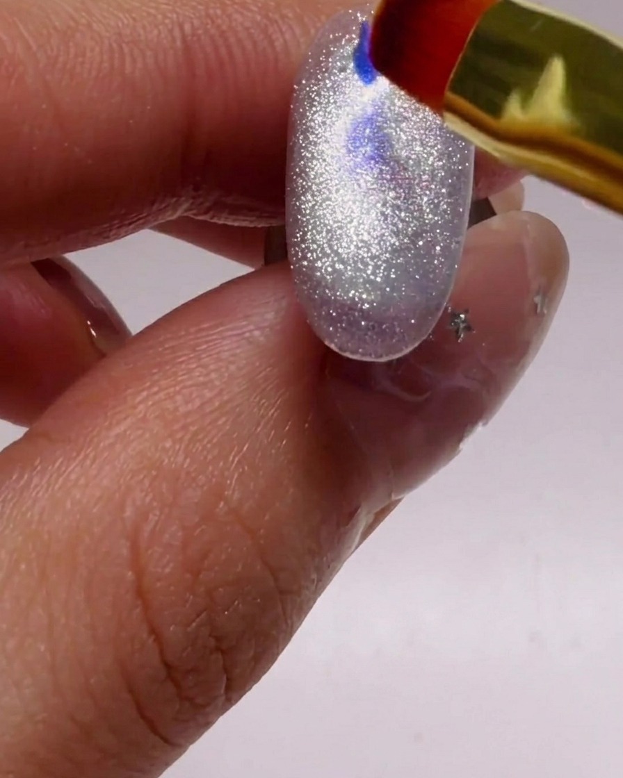 Un pequeño pincel de uñas colocando cuidadosamente hojuelas de glitter iridiscente azul y morado sobre una uña plateada brillante.