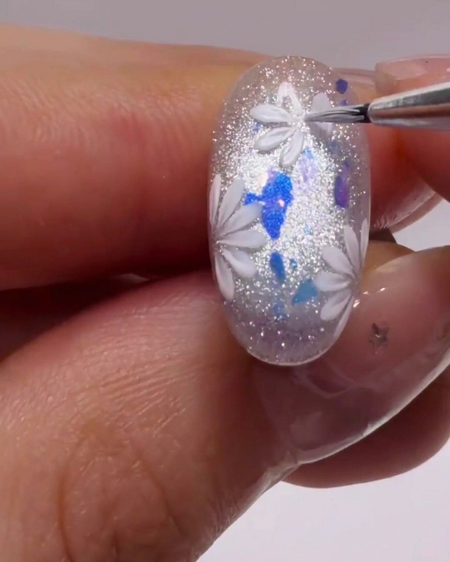 Un pincel delineador fino creando formas precisas de pétalos blancos sobre una uña brillante y con glitter.