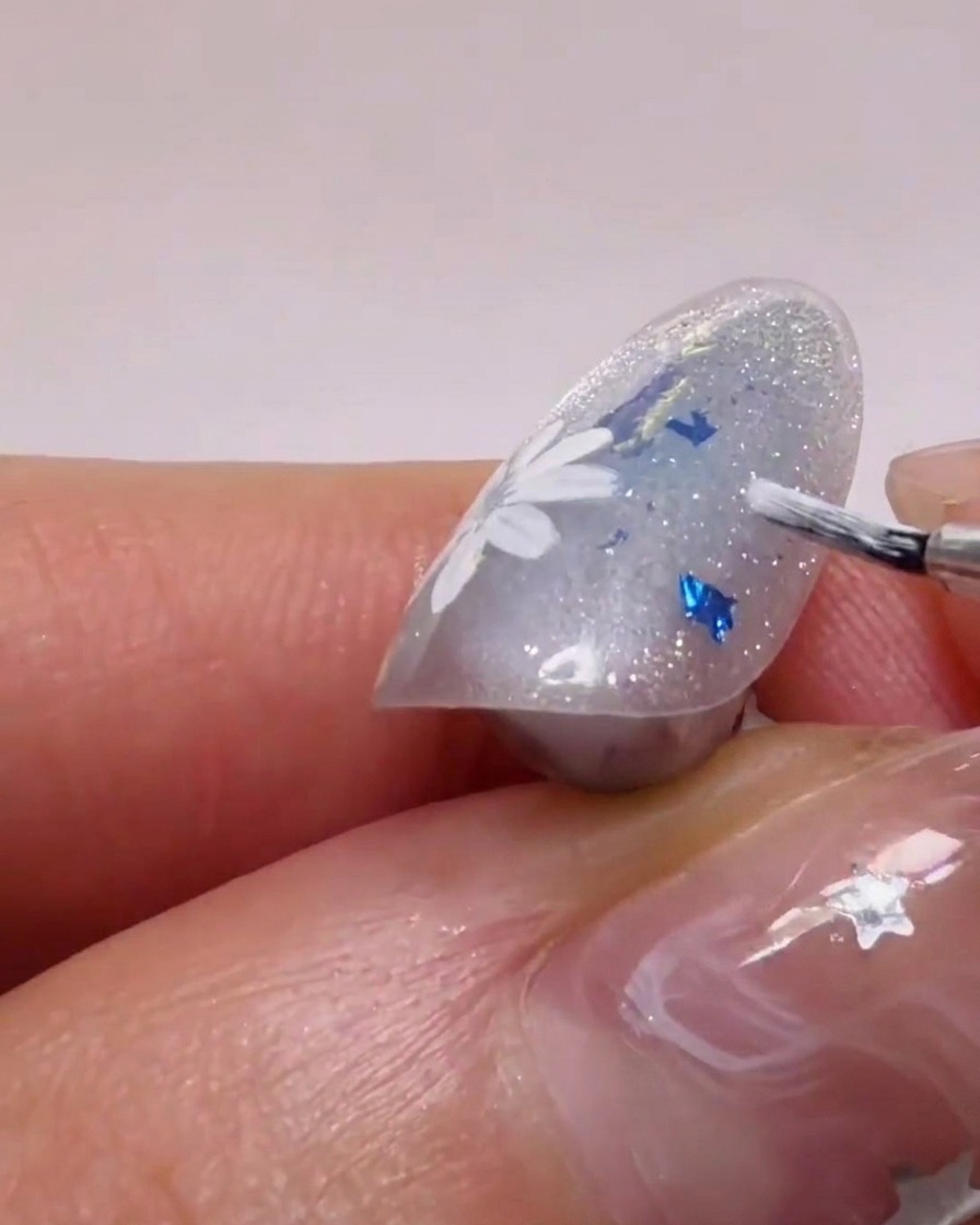 Una vista de cerca de un pincel de uñas aplicando gel blanco para formar pétalos de margarita sobre una base con glitter.