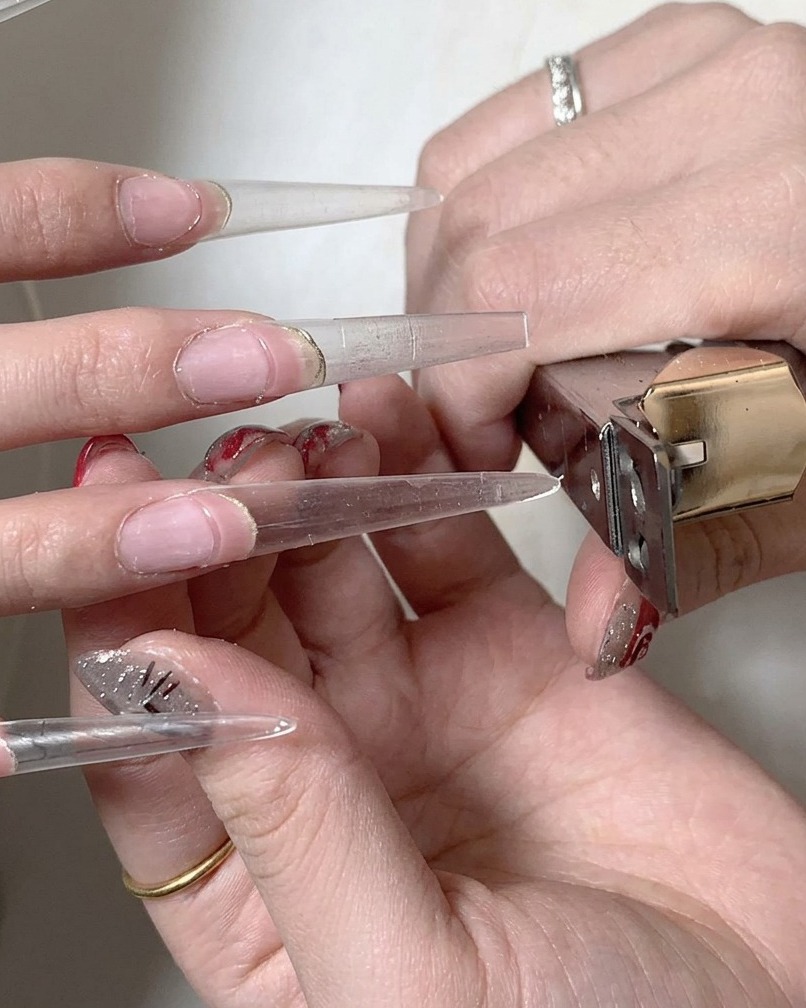 Un pincel aplicando una capa de base coat de gel transparente sobre el punto de unión donde la uña natural se encuentra con la extensión.