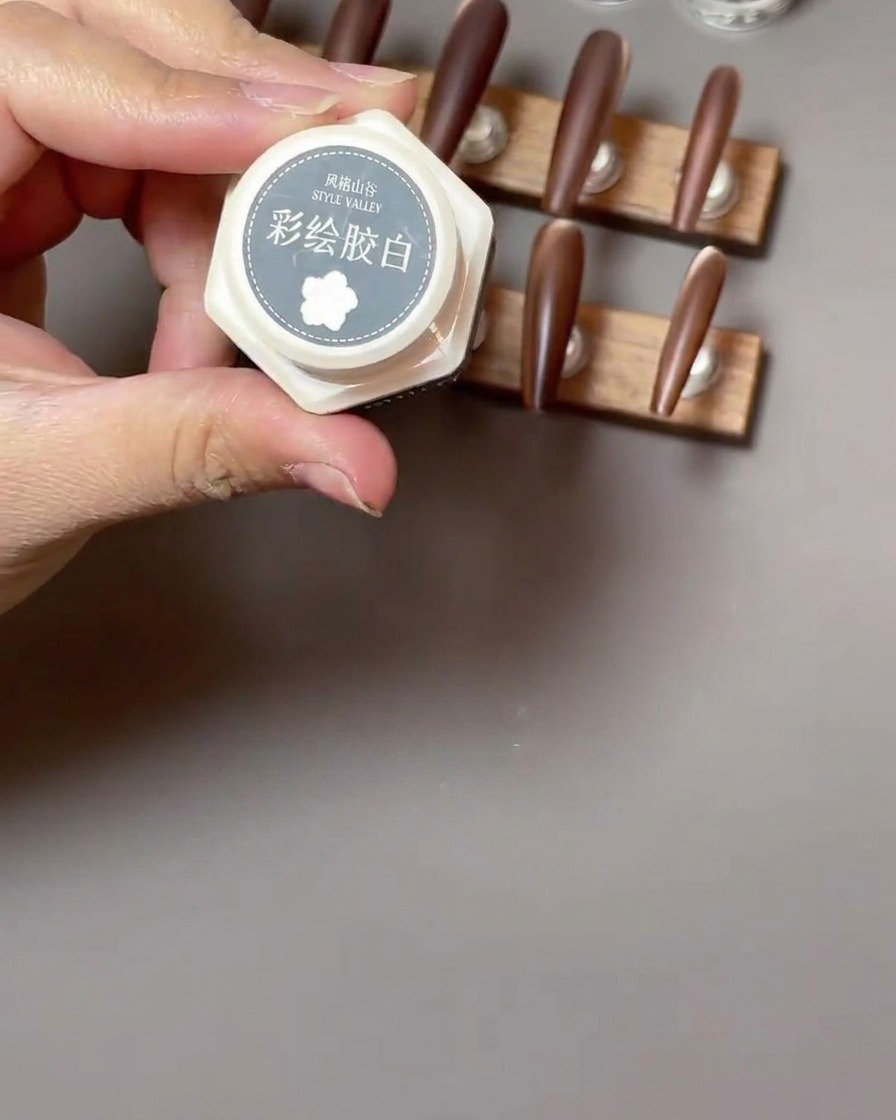 Un pincel delineador fino dibujando pétalos blancos delicados en forma de paréntesis para un diseño de rosa sobre una uña marrón mate.