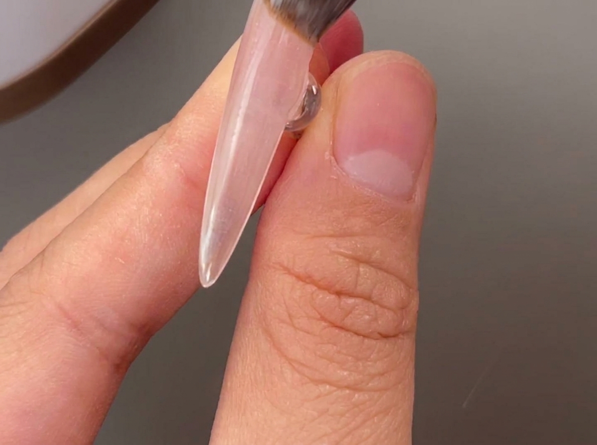 Un pincel aplicando una capa transparente de base gel sobre una uña natural.