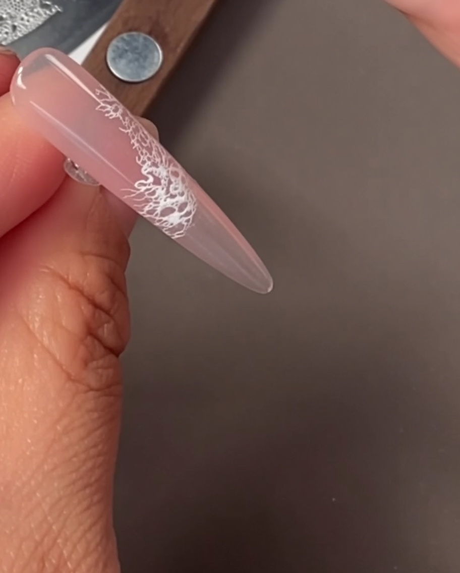 Primer plano de una uña con un delicado patrón de encaje blanco aplicado mediante estampado.