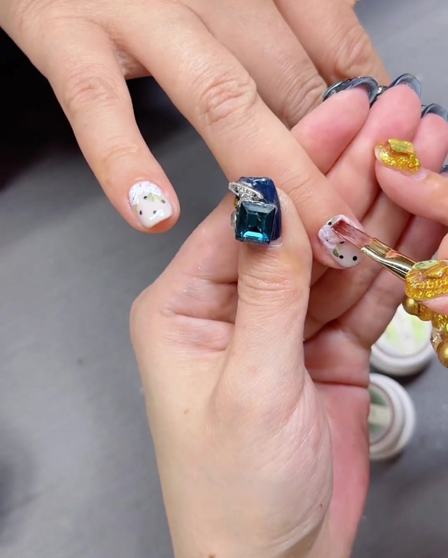 Un pincel aplicando un top coat transparente y brillante sobre una uña con diseño de rosa pintada a mano y lunares.
