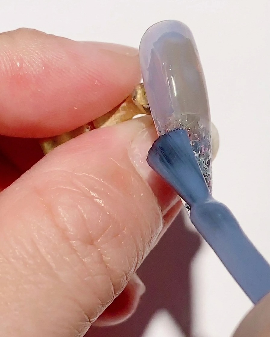 Un pincel de degradado difuminando esmalte de gel azul transparente en la superficie de la uña para una transición suave.