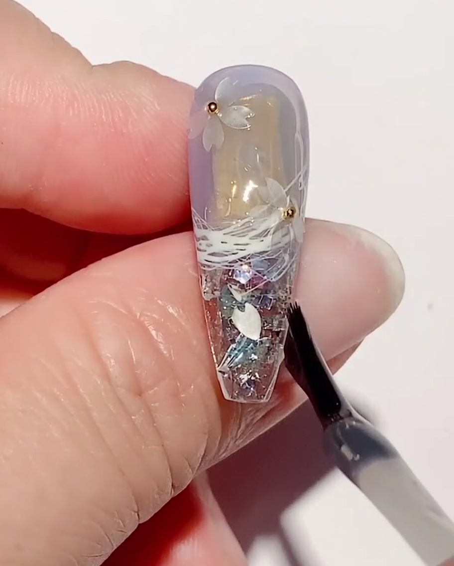 Un pincel de arte de uñas aplicando una capa de gel de refuerzo transparente sobre lentejuelas de flores de cerezo y cuentas de acero en una uña.