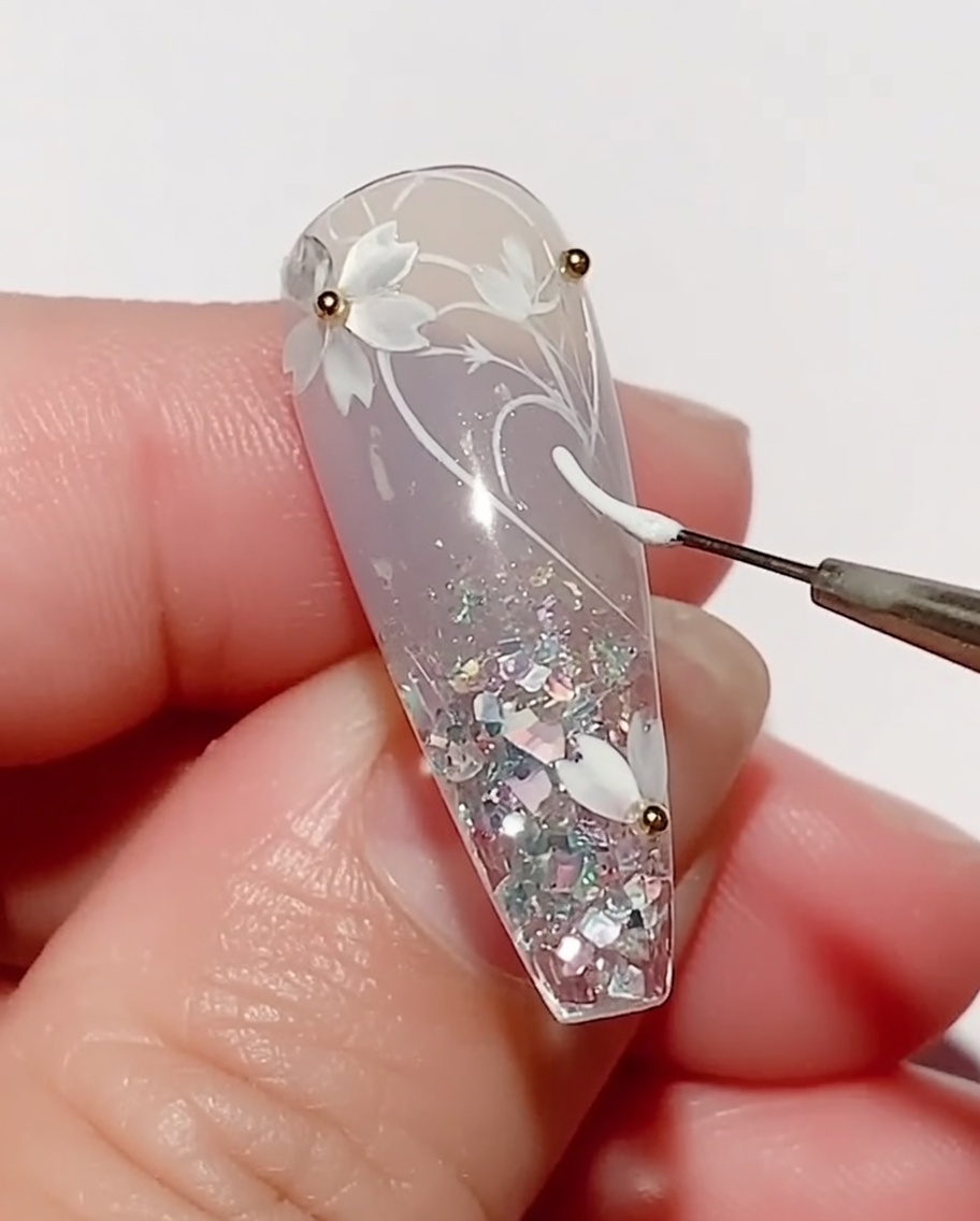 Un pincel fino aplicando gel de color blanco para dibujar la forma de una hoja en una uña decorada con flores de cerezo.